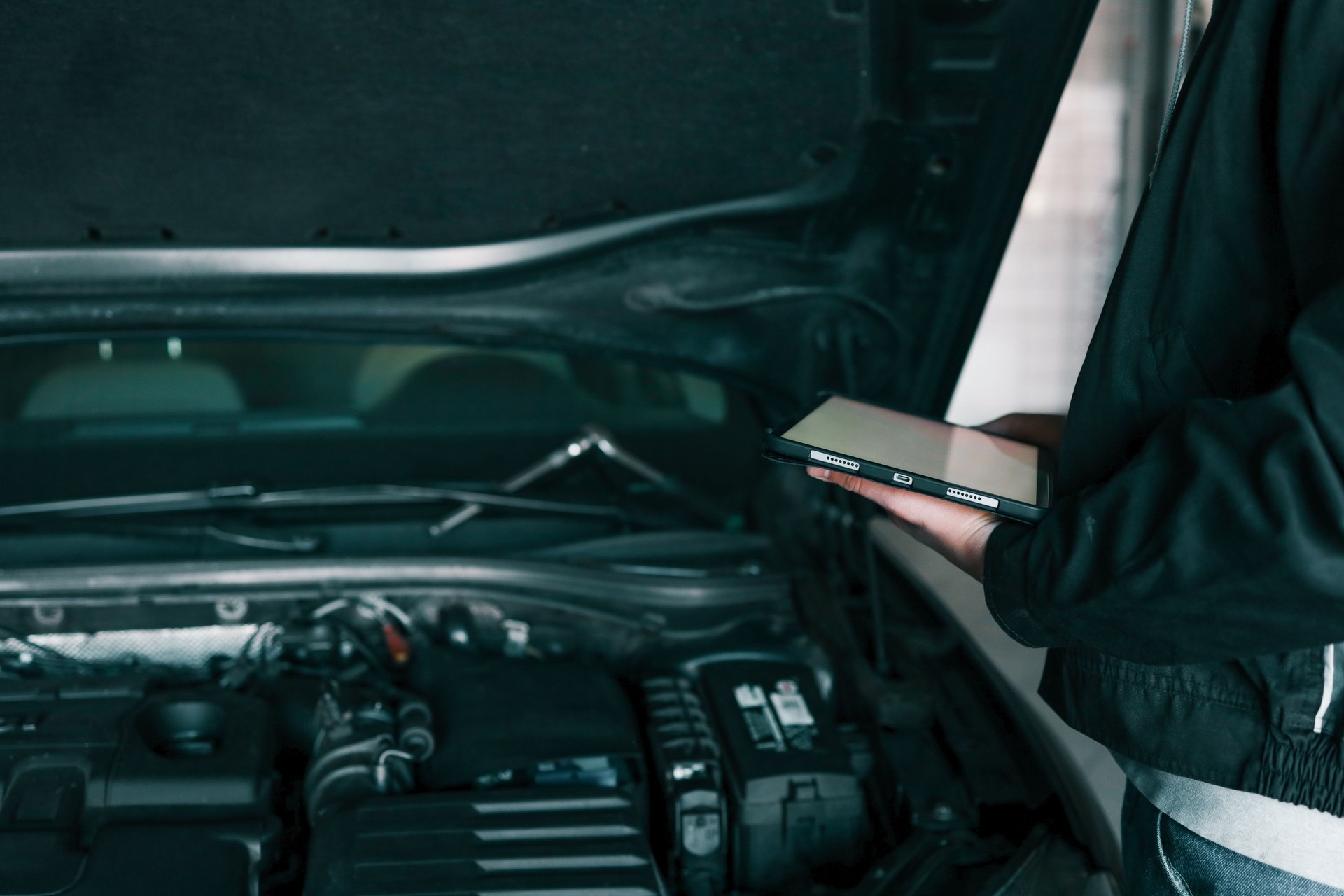 A mechanic holding a tablet while inspecting a car engine in a workshop. The focus is on the integration of modern technology in vehicle diagnostics and repair